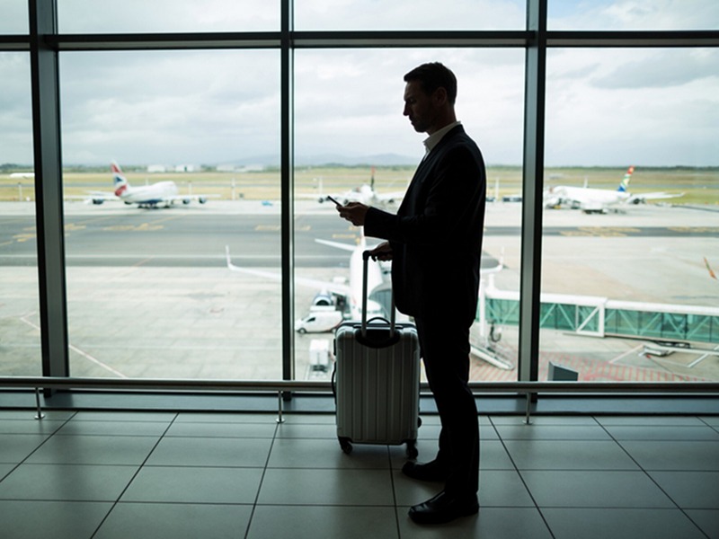 Un homme debout devant une fenêtre d'aéroport, regardant son téléphone et portant une valise 