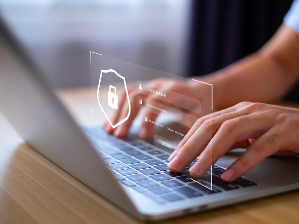 Une image rapprochée montre deux mains d'un utilisateur tapant sur le clavier d'un ordinateur portable argenté. Une interface de connexion virtuelle est affichée au-dessus du clavier. Elle comprend un grand écusson de sécurité blanc avec un cadenas, des champs textuels pour "NOM D'UTILISATEUR" et "MOT DE PASSE" avec des icônes correspondantes, une case à cocher pour "Mémoriser", des liens pour "Mot de passe oublié ?" et "S'inscrire", et un bouton "CONNEXION".